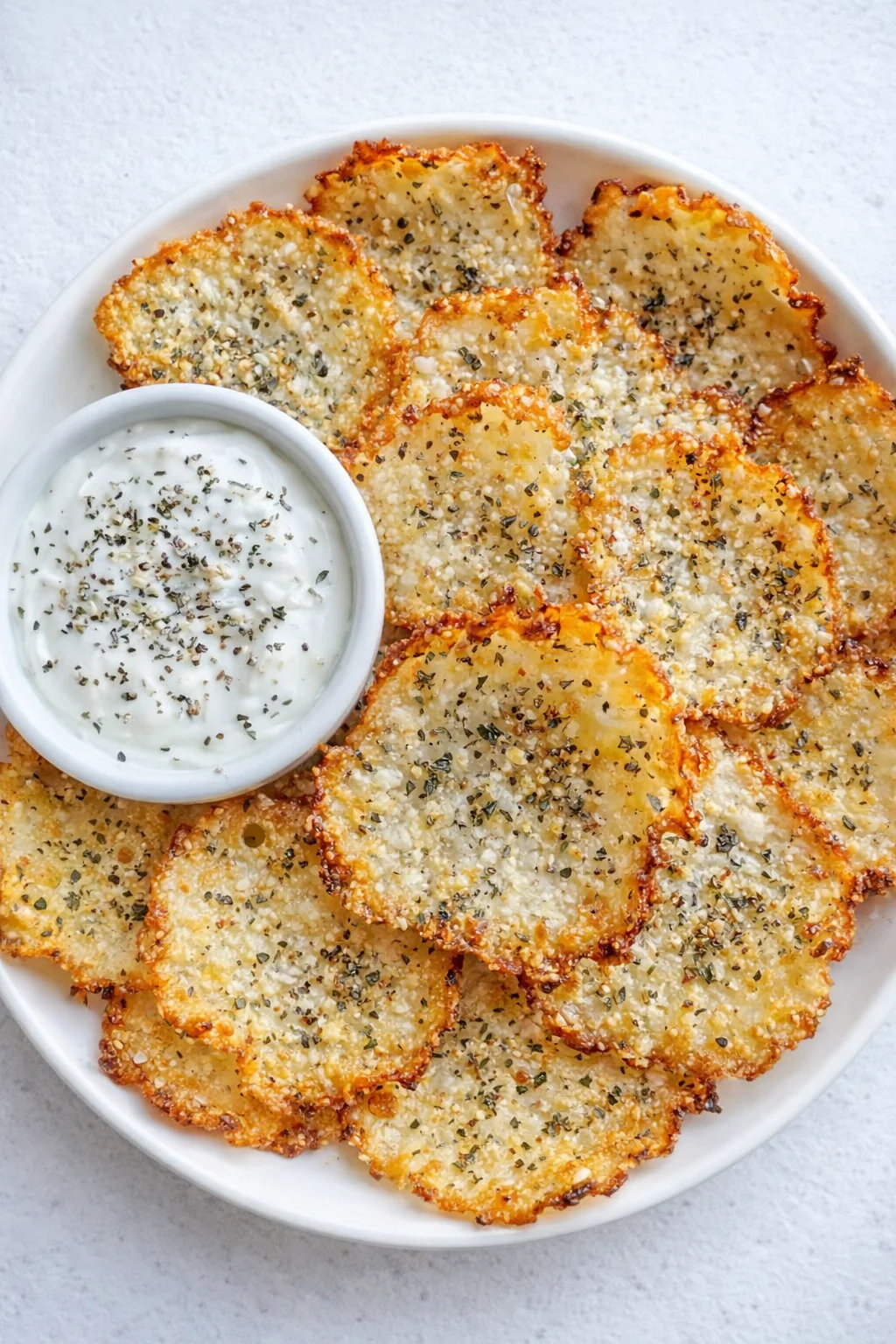Golden-brown potato crisps with herbs on a white plate, served with a creamy herb dip