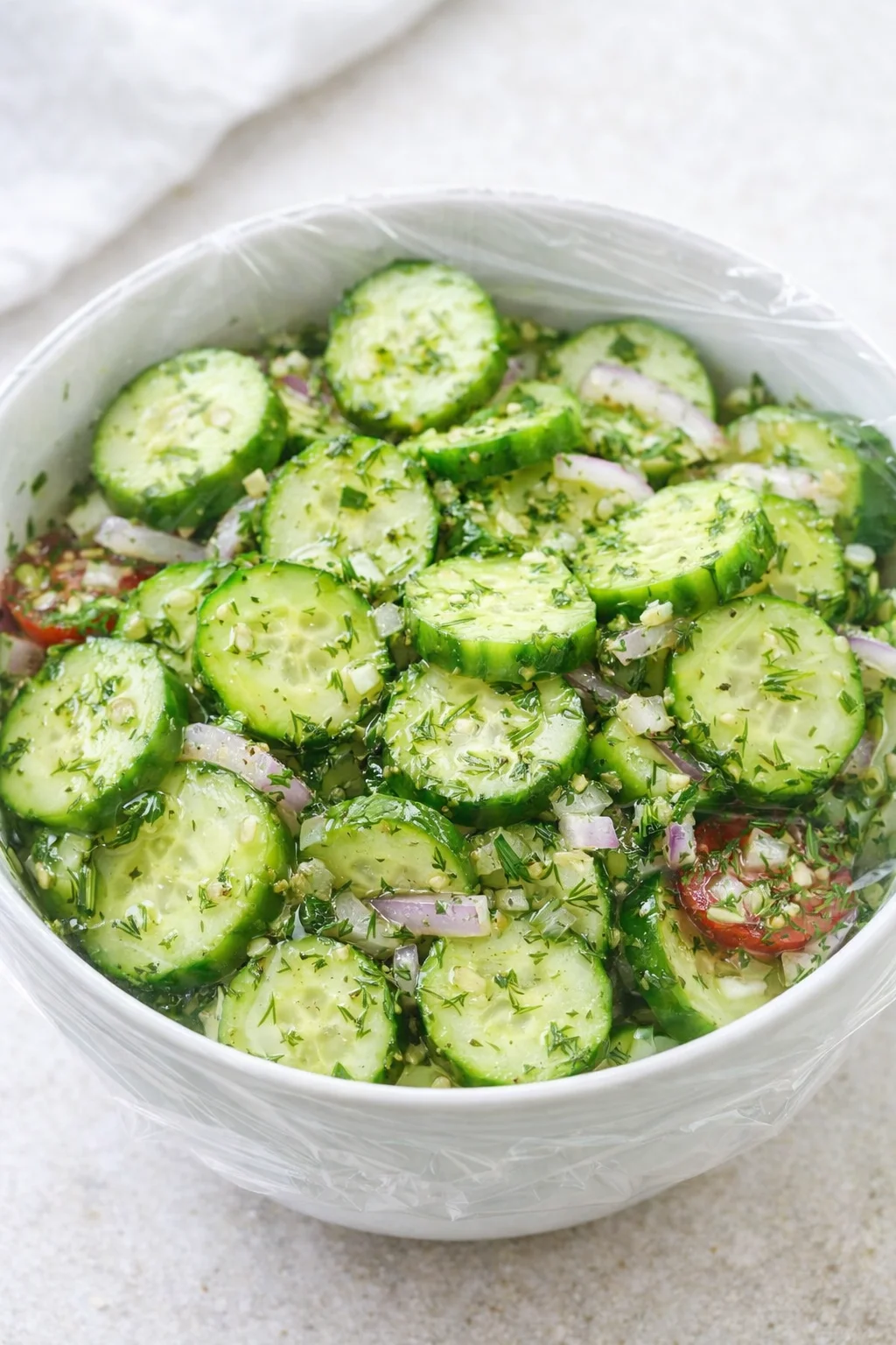 Dressing being poured over cucumber mixture and tossed, chopped dill visible sprinkled on top in bowl.