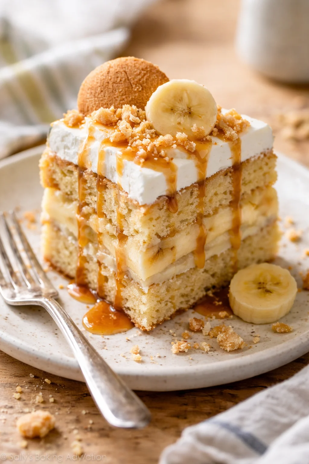 slice of layered banana cake topped with whipped cream, caramel drizzle, banana slices and crumble on a white plate