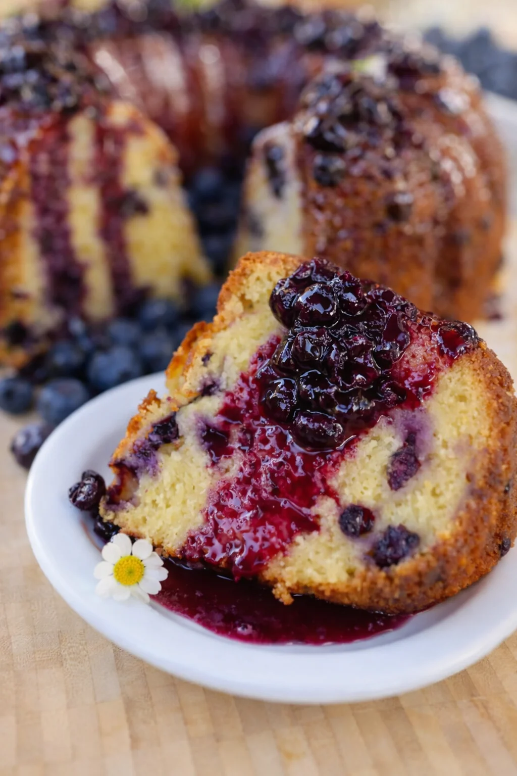slice of blueberry bundt cake with berry glaze on a white plate, whole cake in background