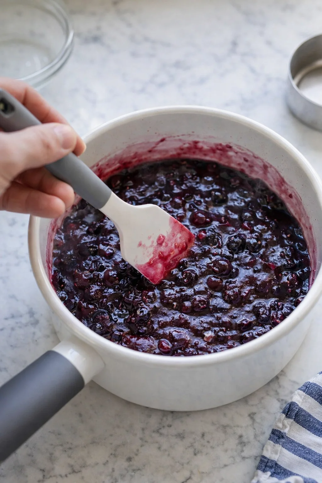 Hand stirring a bubbling blueberry sauce in a white pot on a marble countertop