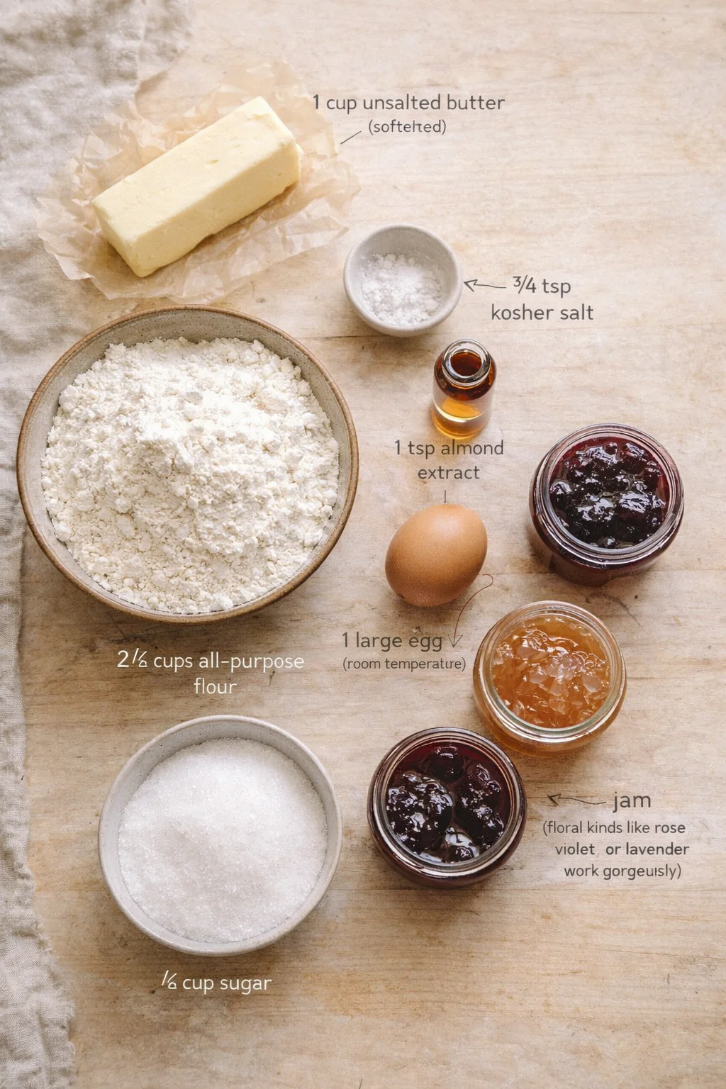 top-down view of baking ingredients on a wooden table: butter, flour, sugar, egg, salt, almond extract, and jam.