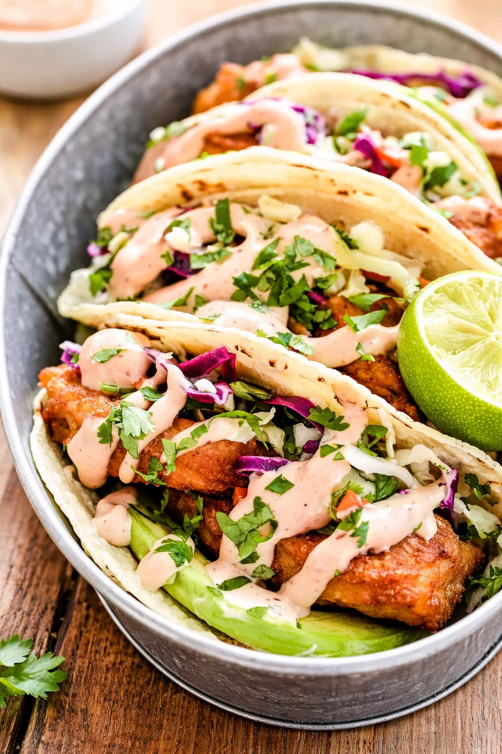 three soft corn tortillas filled with crispy fried chicken, shredded cabbage, cilantro, pink crema, and lime.