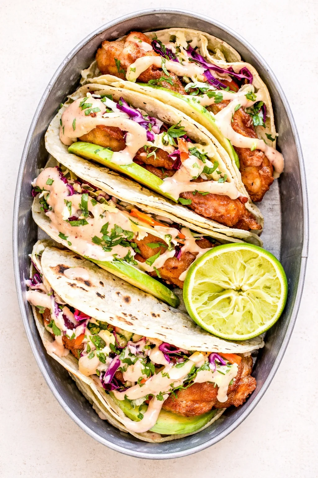 Overhead view of soft tortillas filled with crispy chicken, slaw, avocado, cilantro, and a lime wedge.