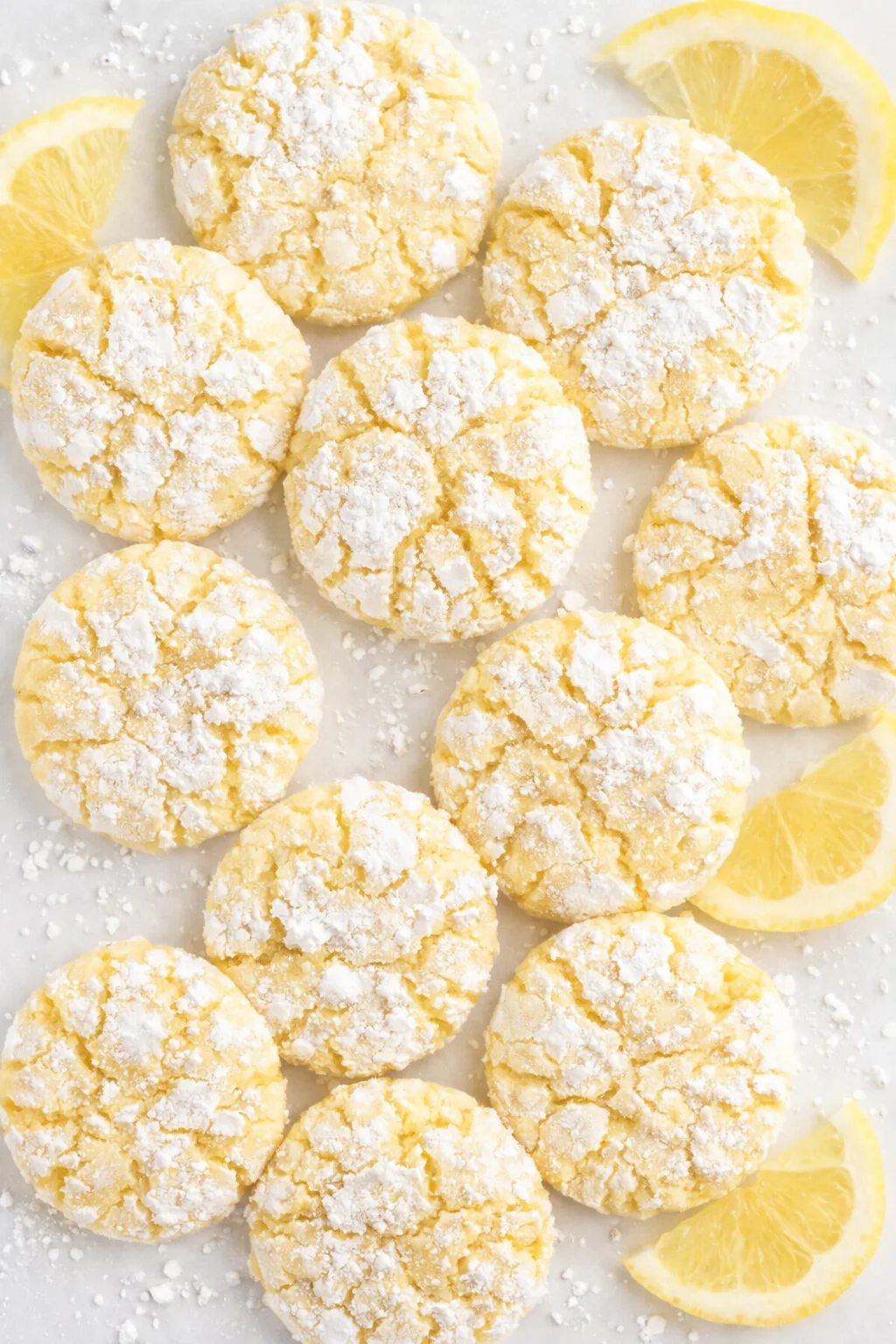 Flat lay of lemon crinkle cookies dusted with powdered sugar, surrounded by lemon wedges on a pale surface.