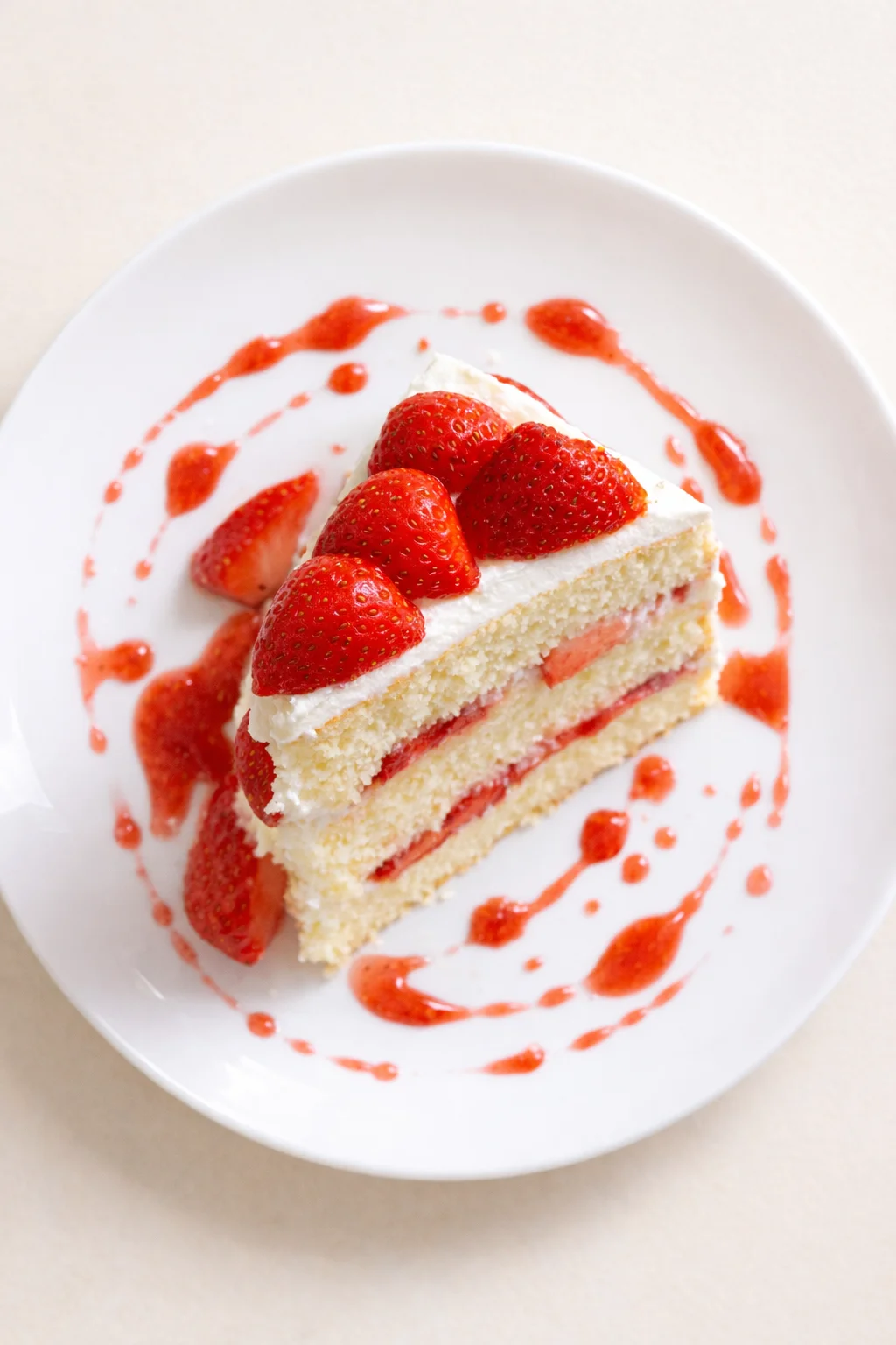 Slice of vanilla cake with strawberry cream, topped with fresh strawberries and red sauce on a white plate.