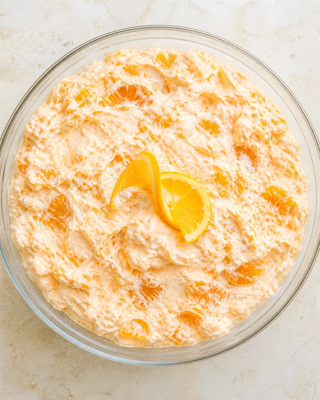 top-down view of a glass bowl containing orange-colored creamy citrus salad with mandarin segments and an orange twist garnish.