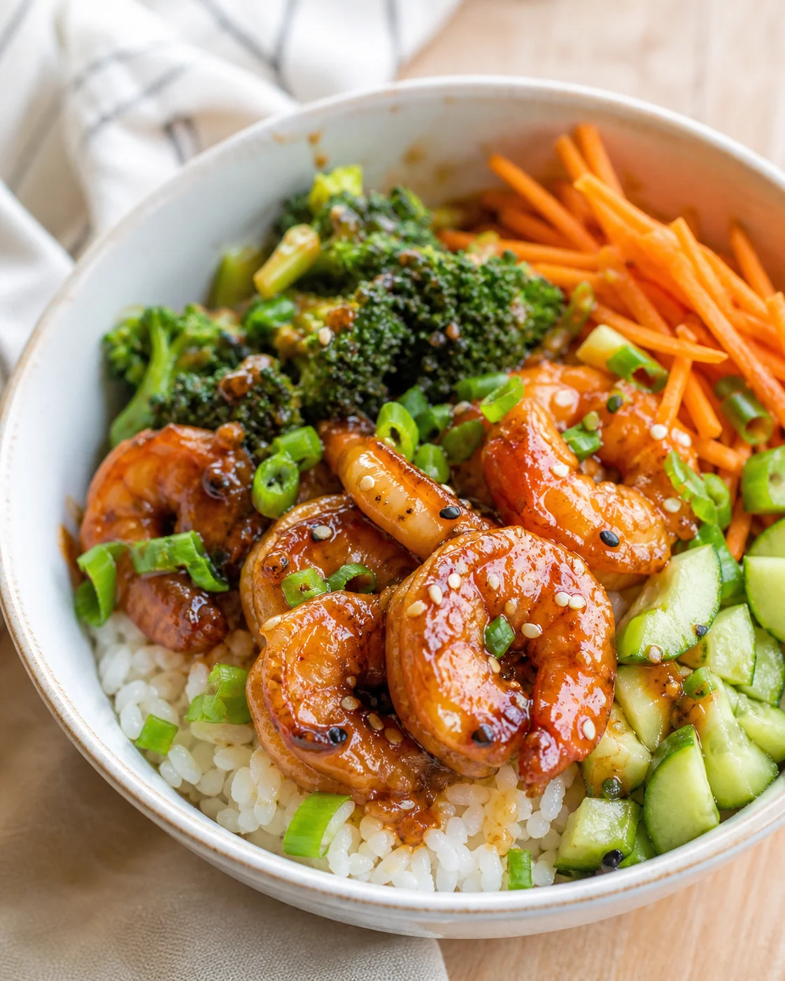 bowl of white rice topped with glazed shrimp, broccoli, shredded carrots, cucumber, green onions, and sesame seeds