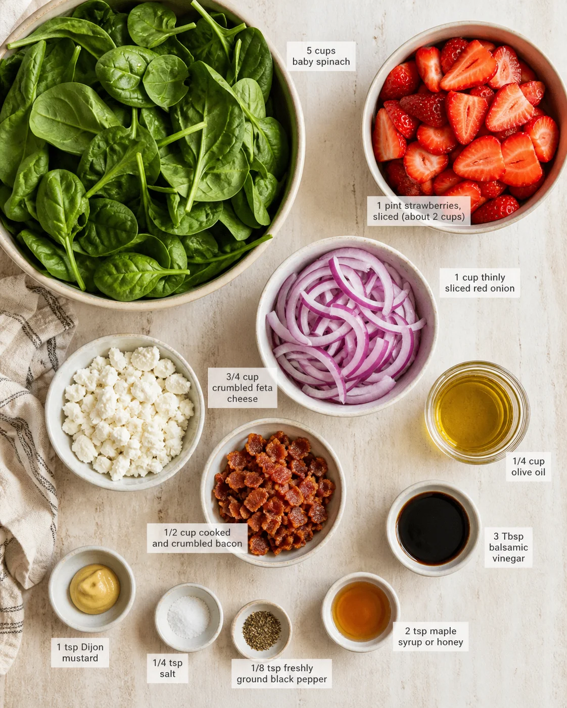 Top-down view of salad ingredients in bowls: spinach, strawberries, onions, feta, bacon, olive oil, balsamic, and seasonings.